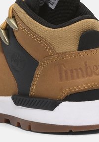 Zapato en tonos marrones y negros, confeccionado con materiales de cuero y textil, que presenta ojales dorados, el logo de Timberland en relieve y una suela de goma blanca.