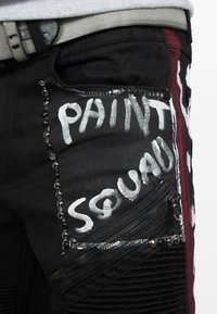 Pantaloni in denim neri con testo dipinto bianco "PAINT SQUAD", tasche con zip, tessuto testurizzato e borchie metalliche lungo le cuciture.