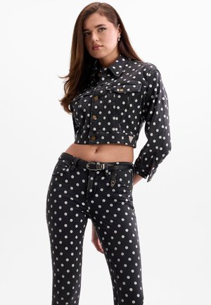 Femme portant une veste en jean courte noire et un jean taille haute à pois blancs, debout avec une main derrière le dos.