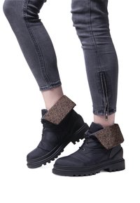 Stivaletti neri alla caviglia con un risvolto in tessuto marrone a motivo, materiale testurizzato e suole in gomma spessa. Indossati con jeans scuri aderenti con cerniere.