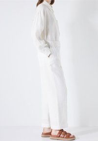 Vit linneskjorta med långa ärmar och en knäppt krage, kombinerad med löst sittande vita linbyxor. Bruna sandaler med remmar lyfter fram stilen.