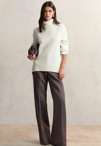 Biely svetrový rolák s rebrovaným lemovaním, skombinovaný s voľnými tmavohnedými nohavicami. Model drží textúrovanú tmavú tašku. Neutrálny pozadie.