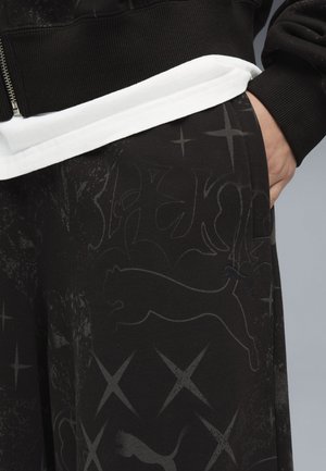 Osoba nosząca czarne spodnie z subtelnym logo Puma i wzorami gwiazd, z ręką w kieszeni, w połączeniu z białą koszulą i czarną kurtką na zamek.