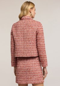 Červený a růžový kostkovaný kabát s límcem, přiléhavý střih a texturovaná tkanina. Nosený s odpovídající sukní, což ukazuje na koordinovaný outfit.