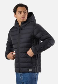 Jeune homme portant une veste matelassée noire à capuche avec des poches zippées, ajustant la poche gauche, debout devant un fond blanc uni.