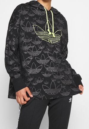 Osoba nosząca czarną bluzę z kapturem Adidas z powtarzającymi się odblaskowymi logo i neonowo żółtymi sznurkami, połączoną z czarnymi spodniami Adidas z białymi paskami.