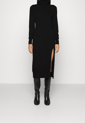 Mujer vestida con un vestido largo negro de cuello alto con una abertura lateral alta y botas negras hasta la rodilla, posando contra un fondo blanco liso.