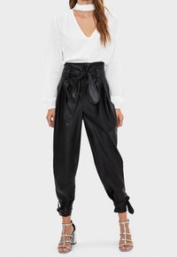 Svarta fusk läderbyxor med hög midja och knytbälte, med en avslappnad passform och samlade ankelmanschetter. Matchas med en vit blus med v-ringning.