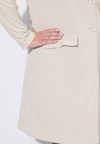 Mano con smalto per unghie rosa chiaro appoggiata sulla patta della tasca di un cappotto beige a costine con bottoni su uno sfondo bianco.