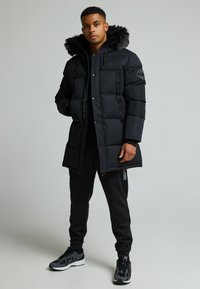 Svart polstret parkas med hette kledd i pels, glidelås og sidelommer. Kombinert med svarte joggebukser og grå løpesko.
