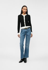 Cardigã preto com acabamento e botões em creme, combinado com jeans de perna reta azul claro e botas pretas de salto.