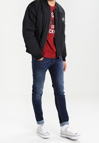 Svart bomberjacka, röd grafisk t-shirt, denimjeans, vita sneakers. Jackan har dragkedjestängning och sidoficka. Jeansen är tapered med uppvikta muddar.