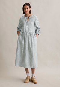 Gestreepte lange jurk in groen en wit katoen, met een keyhole halslijn, driekwartmouwen en zijzakken. Gecombineerd met beige sneakers.