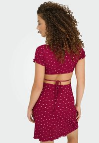 Femme aux cheveux bouclés portant une robe deux pièces à pois rouges avec un dos nu et un détail de lien, se tenant devant un fond clair.