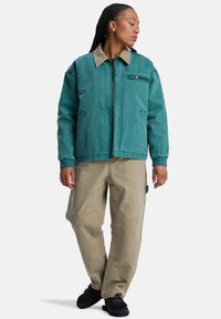 Teal jack met een tan kleurige kraag, ritsluiting en zijzakken, gecombineerd met lichtbeige broeken met een patchworkdetail en een relaxte pasvorm.