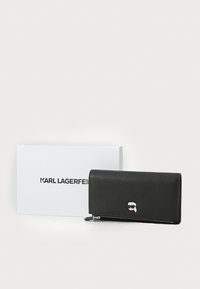 Portefeuille en cuir noir texturé avec fermeture éclair, orné d'un logo argenté. Accompagné d'une boîte blanche de marque.