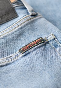 Jasnoniebieska dżinsowa tkanina zbliżeniowo ukazująca czarną metkę z napisem "SUPERDRY" w czerwonym ściegu. Widoczne akcenty szycia i faktura.