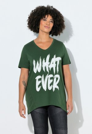 Kiharatukkainen nainen pukeutuneena tummanvihreään V-kaulaiseen T-paitaan, jossa lukee "WHATEVER" suurin valkoisin kirjaimin ja teksturoidut mustat leggingsit.