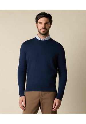 Maglione blu navy a girocollo con un design a maglia liscia, dotato di maniche lunghe e orlo a costine, sovrapposto a una camicia a collare a righe chiare.