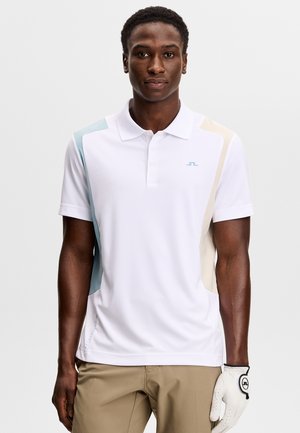 Mężczyzna w białej koszulce polo z jasnoniebieskimi i beżowymi panelami, beżowe spodnie i biała golfowa rękawica na lewej ręce.