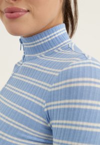 Lyseblå ribbestrikket topp med høy krage og hvite horisontale striper. Har en kort glidelås ved halsen og en tettsittende design.