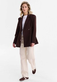 Hnedé sako na mieru s jedným gombíkom, v kombinácii s béžovými širokými nohavicami a tmavohnedými topánkami. Hladká látka a štruktúrovaný dizajn.