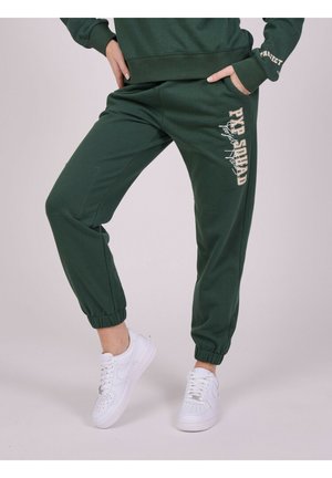 Pantalones de chándal verdes con puños ajustados, con un estampado de 'PYP SQUAD' en crema. Combinados con zapatillas blancas para un look casual.