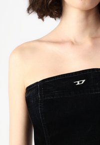 Černý denimový top bez ramínek se strukturovaným designem, který má stříbrné logo s kovovými doplňky a kontrastní švy podél okrajů.