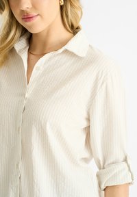 Crème button-up shirt met een subtiel verticaal streepjespatroon, opgerolde mouwen en een zachte, textuurstof.