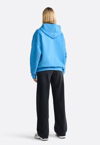 Sudadera azul oversized con cordón, puños y dobladillo acanalados, combinada con pantalones anchos negros y zapatillas blancas. Vista trasera.