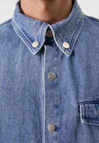 Camisa de denim azul claro com colarinho clássico, botões de pressão prateados e um bolso no peito com uma textura de borda desfiada.