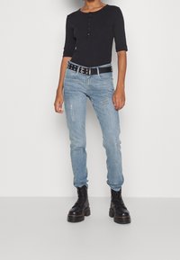 Top aderente nero con maniche tre quarti, jeans blu strappati a vestibilità slim e stivali neri con lacci e suola spessa. Cintura con fibbia argentata.