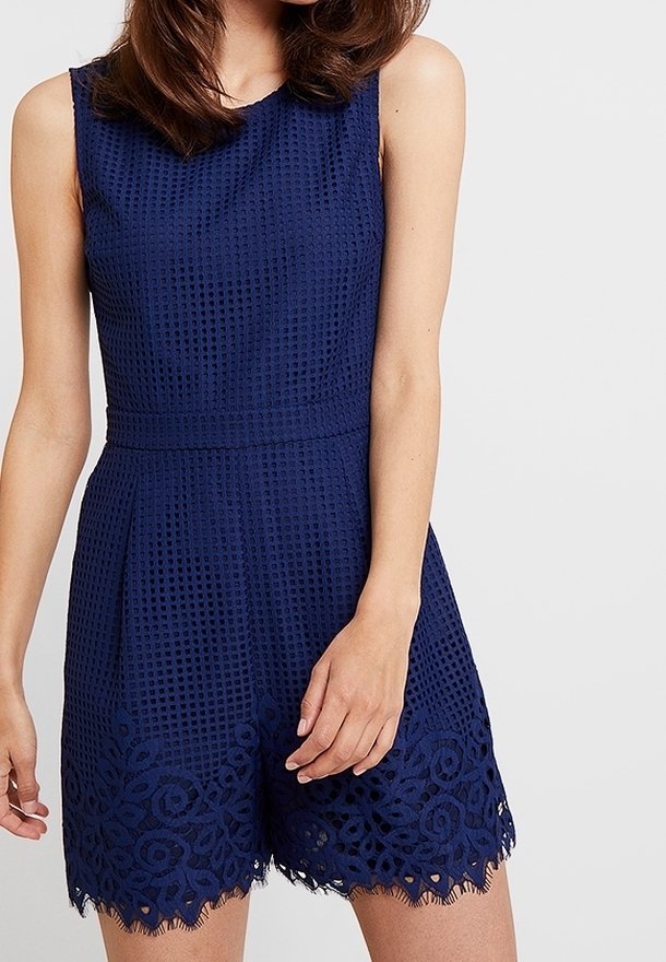 Bezrukávový romper v námořnické modři s texturovanou látkou a květinovou krajkovou lemovkou, nošený částečně viditelnou osobou na bílém pozadí.