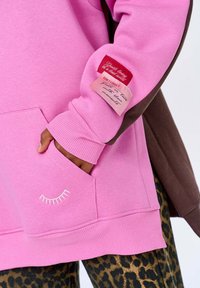 Růžová mikina s přední kapsou, zdobená bílou výšivkou řas a látkovými štítky v červené a růžové na hněvém rukávu.