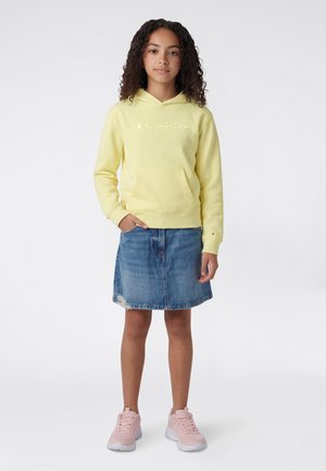 Gelber Kapuzenpullover mit Fronttasche und gesticktem Logo, kombiniert mit einem Jeansrock und rosa Sportschuhen.