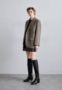 Blazer de lana en espiga marrón con un solo botón, combinado con una minifalda negra y botas de cuero negras hasta la rodilla con un acento dorado.