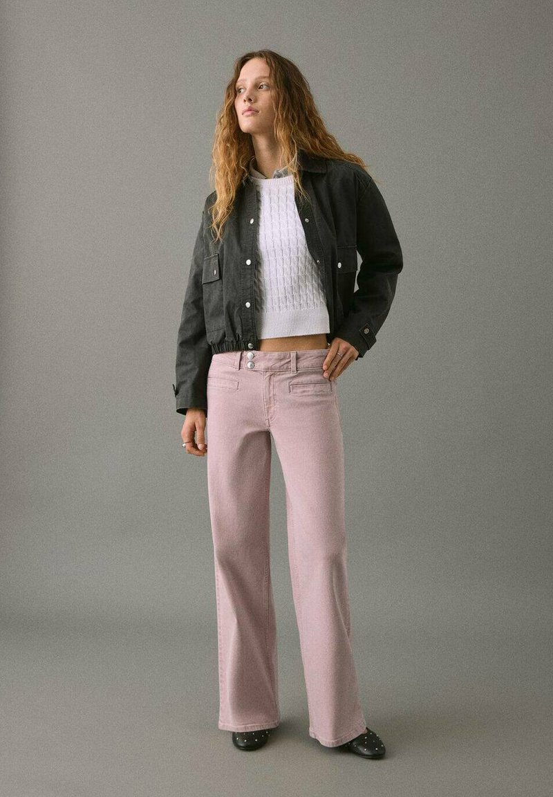 Chaqueta negra corta con bolsillos frontales, suéter blanco texturizado, pantalones de pierna ancha rosa claro y zapatos negros con acentos.