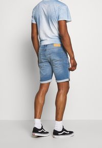 Man som bär ljusblå uppvikta denimshorts, en ljusblå urblekt t-shirt, vita strumpor och svarta sneakers, stående mot en enfärgad bakgrund.