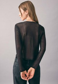 Eine Frau mit langen Haaren, die ein durchscheinendes schwarzes Langarmoberteil und schwarze Jeans trägt, steht mit hinter dem Rücken verschränkten Händen.
