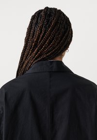 Svart jakke med et uformelt design, som har spiss krage og synlig sømdetaljering. Bak viser det fletet hår som faller ned.