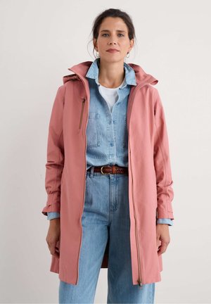 Femme portant un manteau à capuche rose poudré par-dessus une chemise en denim bleu clair et un jean, avec une ceinture tissée marron, se tenant devant un fond uni.