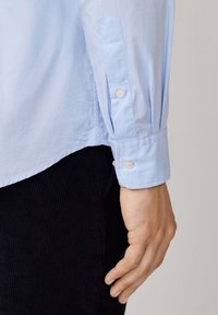 Camisa de vestir azul claro con una textura suave, puños abotonados y detalles de costura sutiles, combinada con pantalones de pana oscuros.