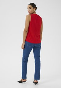 Frau in einem roten ärmellosen, gemusterten Oberteil, blauen Jeans und schwarzen High Heels, die steht und über die Schulter schaut, vor einem einfarbigen Hintergrund.