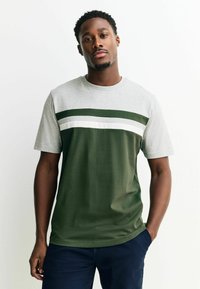 Man met kort krullend haar, gekleed in een t-shirt met kleurvlakken en horizontale strepen in groen, wit en grijs, staat met één hand in zijn zak.