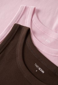 Gros plan de débardeurs en tissu côtelé rose et marron, montrant des encolures rondes et des étiquettes de taille « TWO SOON » à l'intérieur des cols.