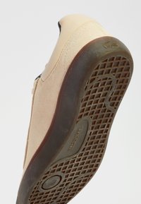 Baskets en daim beige avec une semelle en caoutchouc marron foncé montrant un motif de grip texturé et le logo « Lacoste » au centre de la semelle.