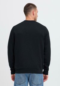 Sudadera negra hecha de un tejido suave, con cuello redondo y mangas largas, con un dobladillo y puños acanalados, vista desde la parte de atrás.
