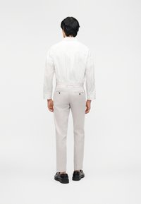 Άνδρας που στέκεται με την πλάτη, φοράει ανοιχτό μπεζ παντελόνι με πίσω τσέπες και λευκό πουκάμισο με μακριά μανίκια και διακριτικές κάθετες ρίγες.