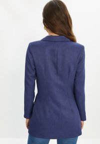 Blazer bleu marine ajusté en tissu texturé, avec col cranté, manches longues et silhouette structurée. Vue de dos.