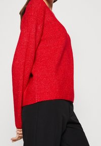Pull en tricot rouge à coupe décontractée et décolleté en V. Tissu texturé avec une légère brillance. Porté avec un pantalon noir, les mains posées sur les côtés.